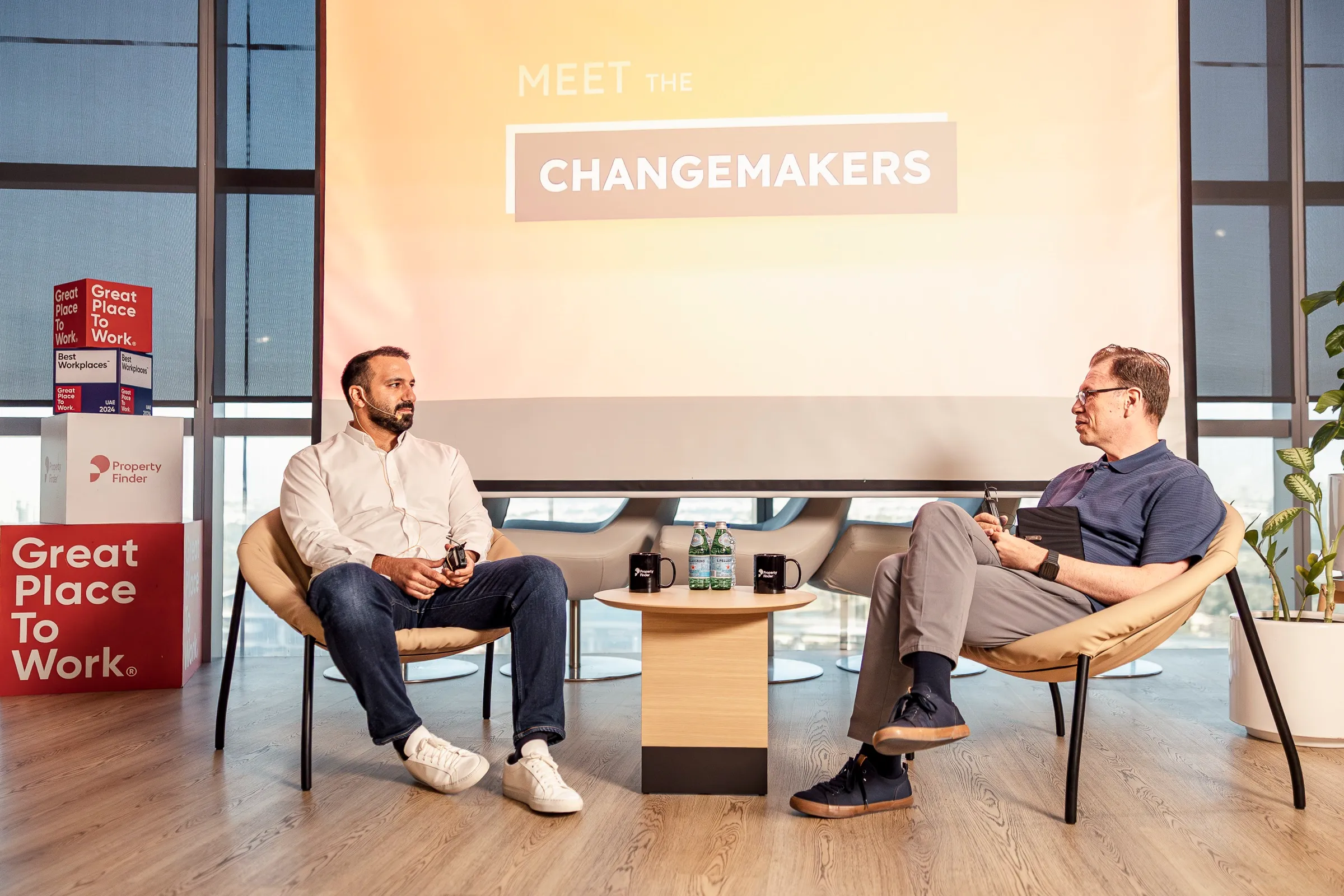 Two speakers in a fireside-format conversation on a bright stage set in front of a projected title